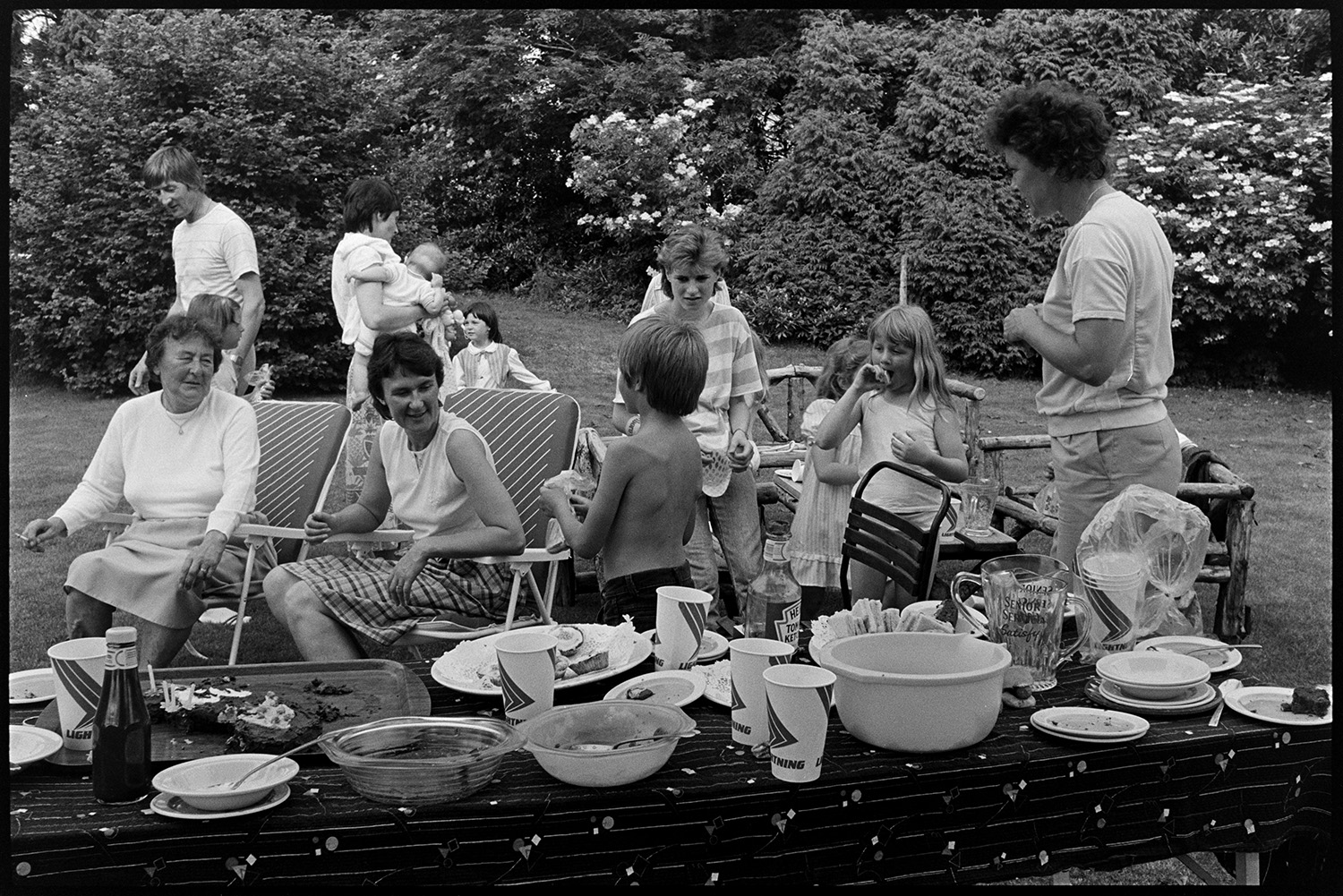Barbeque in garden. 
[A man, women and children sat on garden chairs and benches, and eating barbequed food in the garden at Barlands, Dolton. A table laid with food and drinks can be seen in the foreground.]
