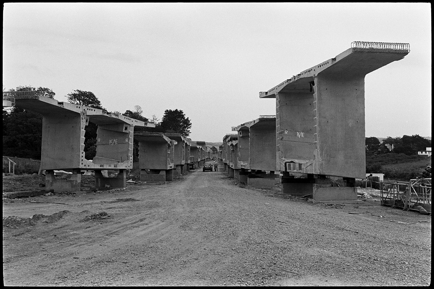 Sections of bridge before assembly, near the river. Concrete. 
[Concrete sections of Bideford New Bridge lined up in two rows on a track near to the River Torridge, before being assembled across the river. A car can be seen between the two rows.]