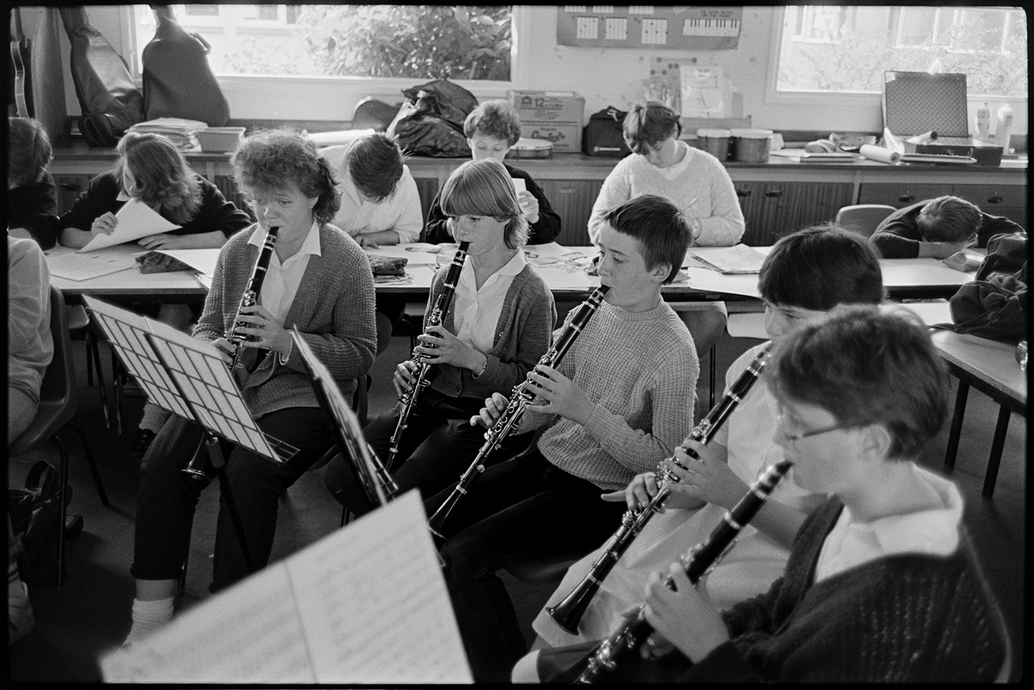 Music class in school, visiting teacher with staff and pupils playing music and advising. 
[A group of boys and girls playing clarinets in a classroom at Holsworthy School. Other pupils are working on desks in the background.]