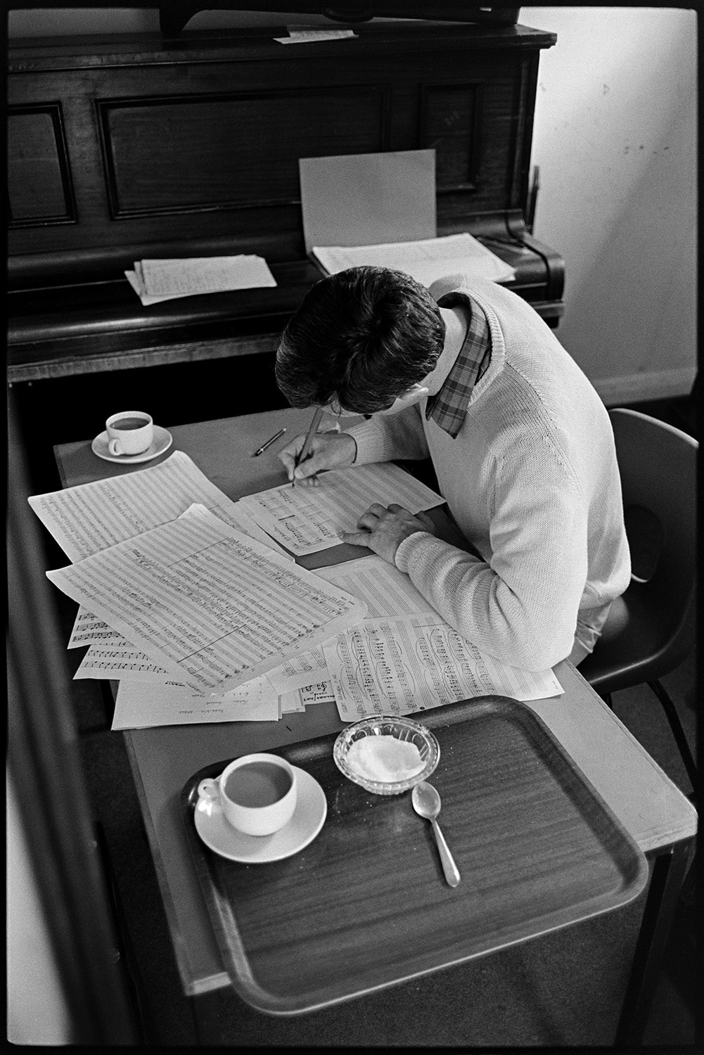 Music class in school, visiting teacher with staff and pupils playing music and advising. 
[Chris Williams writing out music on a desk at Holsworthy School. A tray with a cup of tea and bowl of sugar are beside him. An upright piano can be seen next to the desk.]