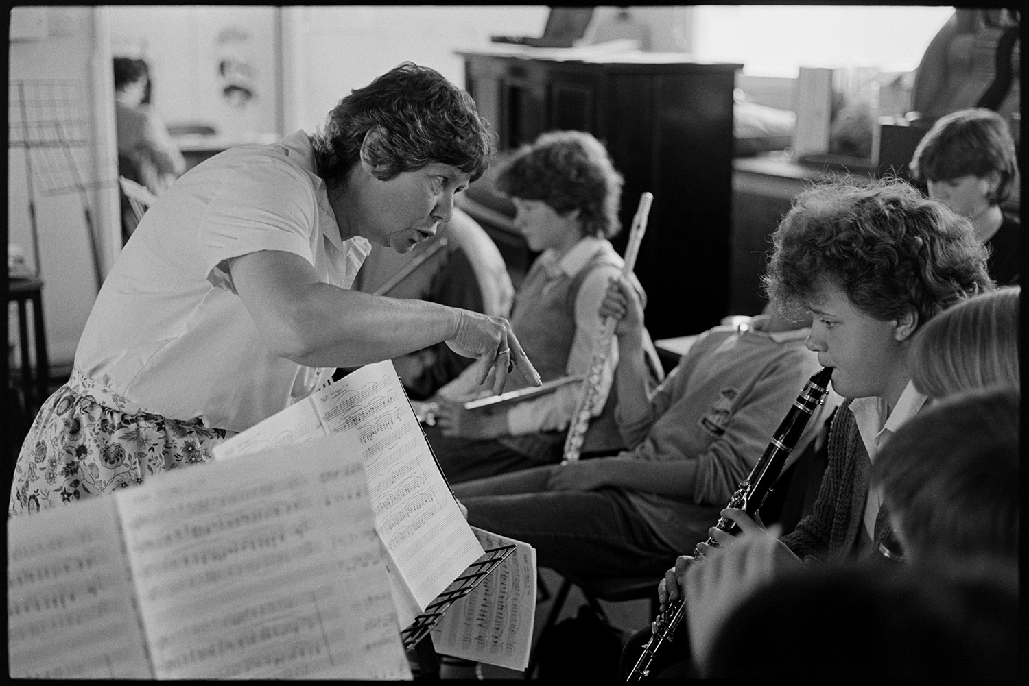 Music class in school, visiting teacher with staff and pupils playing music and advising. 
[A woman teaching a music lesson or orchestra in Holsworthy School. She is talking to a child playing the clarinet. Pupils with flutes can be seen in the background.]