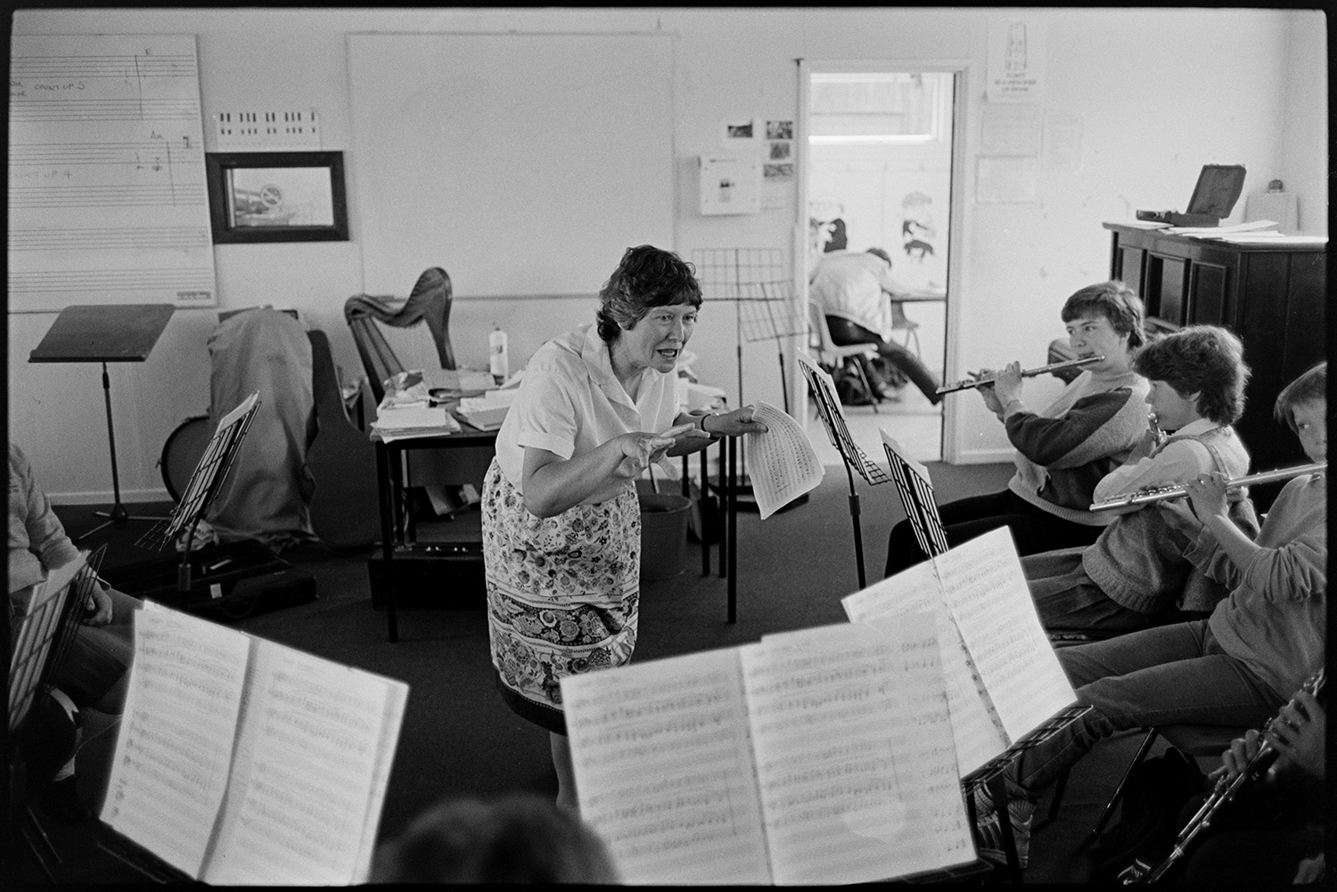 Music class in school, visiting teacher with staff and pupils playing music and advising. 
[A woman teaching a music lesson or orchestra in Holsworthy School to pupils playing flutes and clarinets. A harp can be seen behind the desk in the background.]