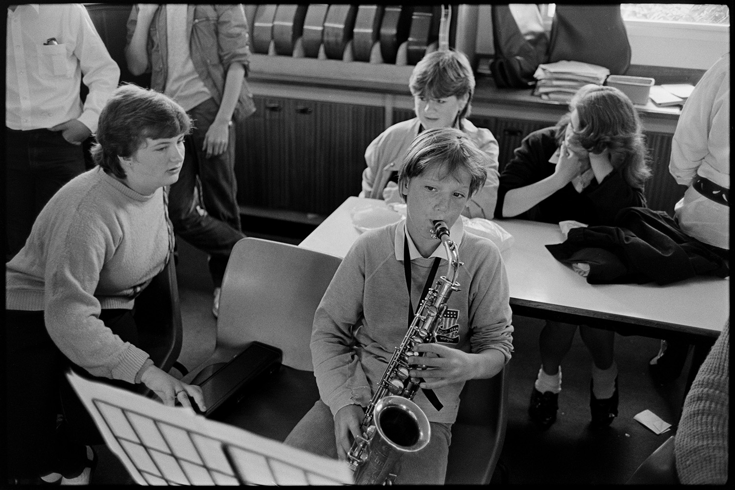 Music class in school, visiting teacher with staff and pupils playing music and advising. 
[A boy playing a saxophone in a music lesson or orchestra practice at Holsworthy School. Other children are watching and listening in the background.]