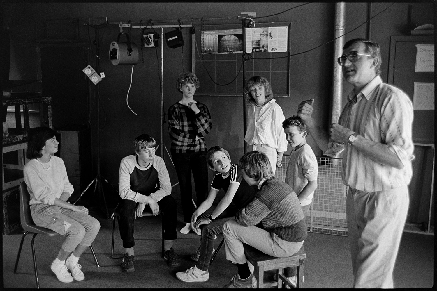Music class in school, visiting teacher with staff and pupils playing music and advising. 
[A man teaching a drama class at Holsworthy School. Lighting equipment can be seen in the background.]