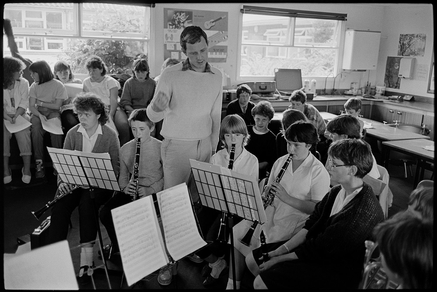 Music class in school, visiting teacher with staff and pupils playing music and advising. 
[Chris Williams teaching a music lesson in Holsworthy School. He is teaching a group of children playing clarinets. More children are sat in the background, possibly singing with the school orchestra.]