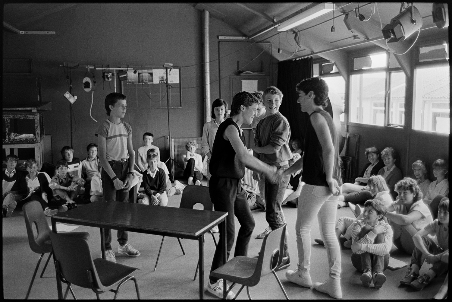 Music class in school, visiting teacher with staff and pupils playing music and advising. 
[Pupils in a drama class at Holsworthy School. Six children are acting by a desk and chairs while the rest of the class watches. Lighting equipment is hung up around the drama studio.]