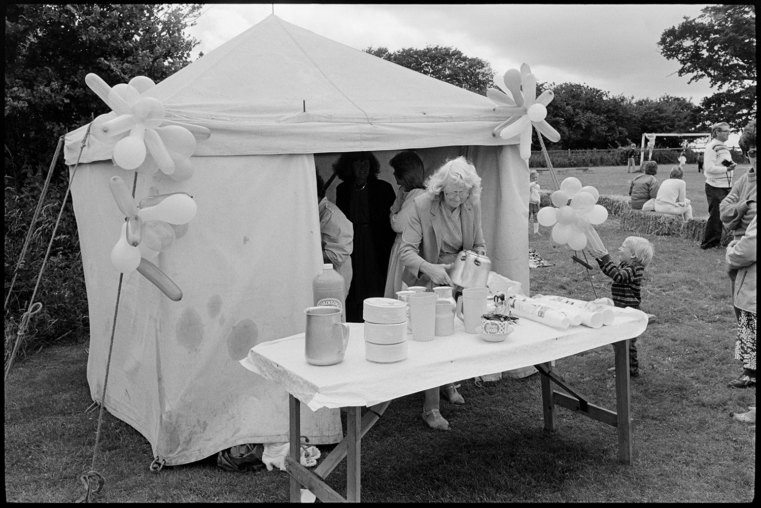 School sports day, refreshment tent, children and spectators. 
[A woman setting up refreshments on a table outside a small tent with balloons at Dolton School sports day. Cups, a teapot, jugs and bottle of Robinsons squash are on the table. A young child is looking at the balloons on the tent and people are watching sports in the background.]
