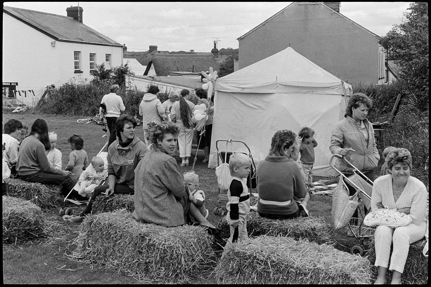 School sports day, refreshment tent, children and spectators. 
[Women and children sat on hay bales at Dolton School sports day. Some of the women have prams. People are gathered around a refreshment table in the background by a small tent.]