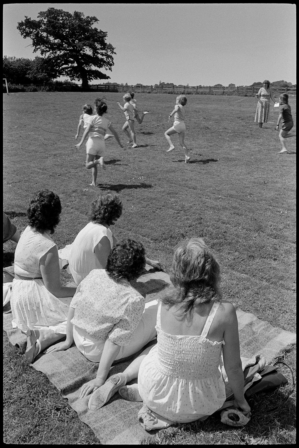 School sports day, races, people sitting in sun and shade waiting to run. 
[Four women sat on picnic rugs at Dolton School sports day. They are watching children compete in a running race.]