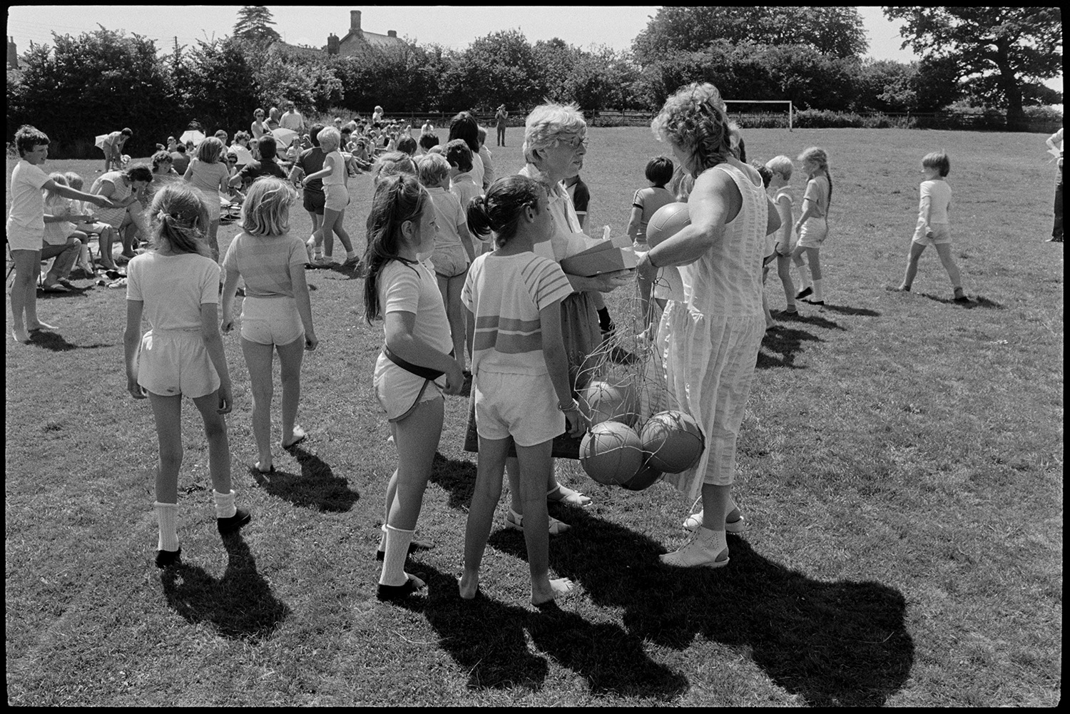 School sports day, races, people sitting in sun and shade waiting to run. 
[Children getting ready for races at Dolton School sports day. A teacher is holding a bag of balls and talking to another woman. Spectators are sitting along the side of the track in the sports field.]