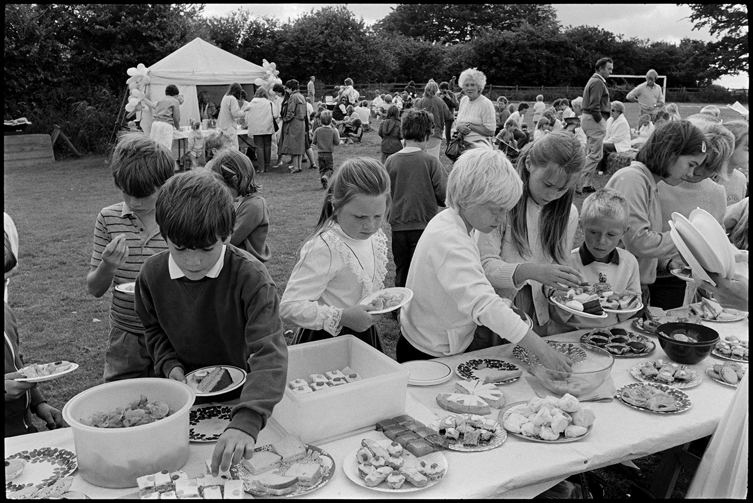 School sports day, races ,people having tea and cakes from trestle tables. 
[Children helping themselves to cakes and crisps laid out on trestle tables at Dolton School sports day. Other people can be seen in the background on the sports field and at a refreshment tent with balloons.]