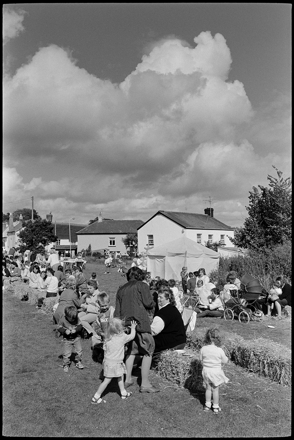 School sports day, races ,people having tea and cakes from trestle tables. 
[Parents and children sat on hay bales having refreshments at Dolton School sports day. Some of the women have prams and a refreshment tent can be seen in the background.]
