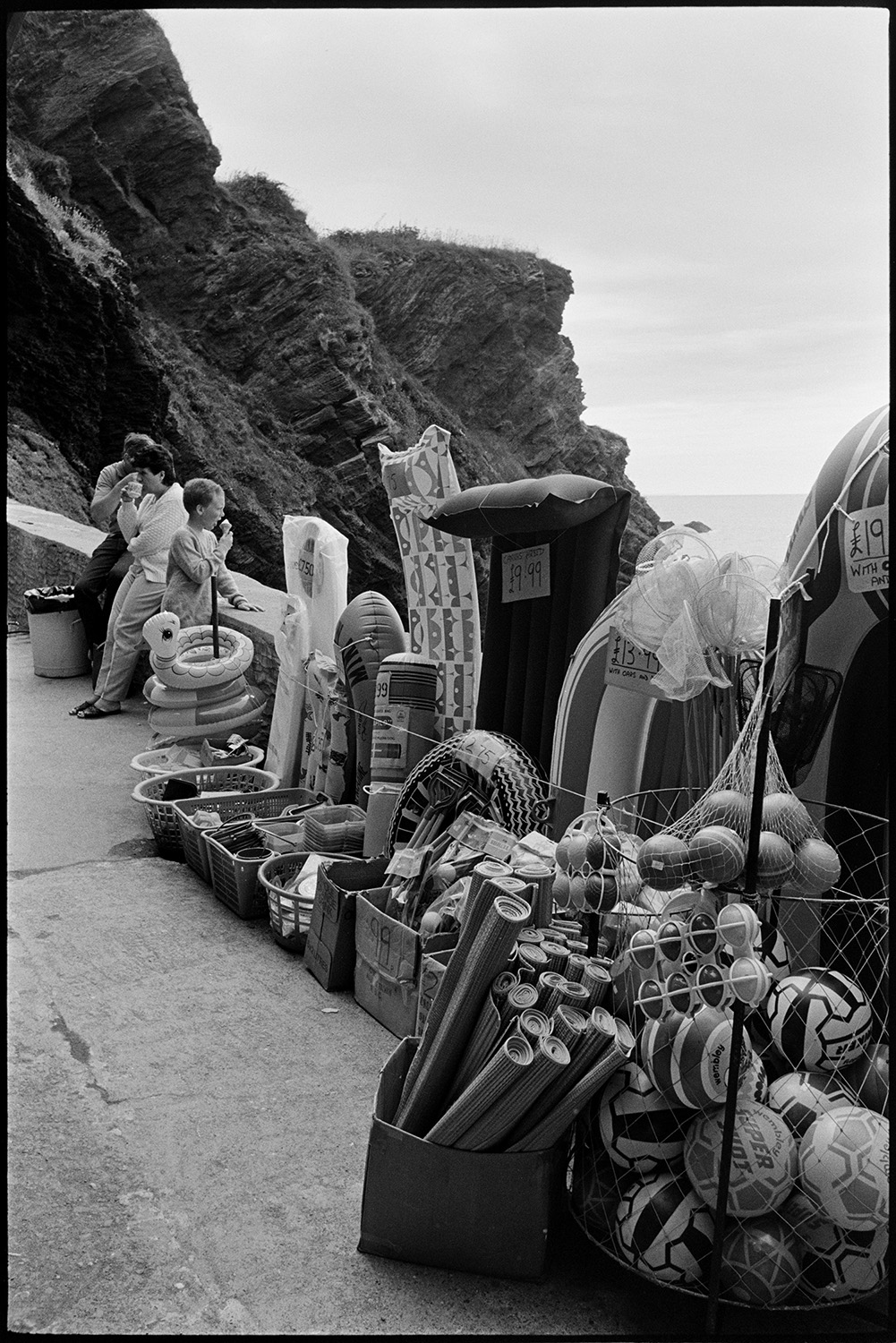 Seaside goods on display. 
[Beach goods, including footballs, rubber rings, inflatable dinghys and beach mats, on display by the sea wall in Ilfracombe. People eating ice creams and drinking tea can be seen in the background by cliffs.]