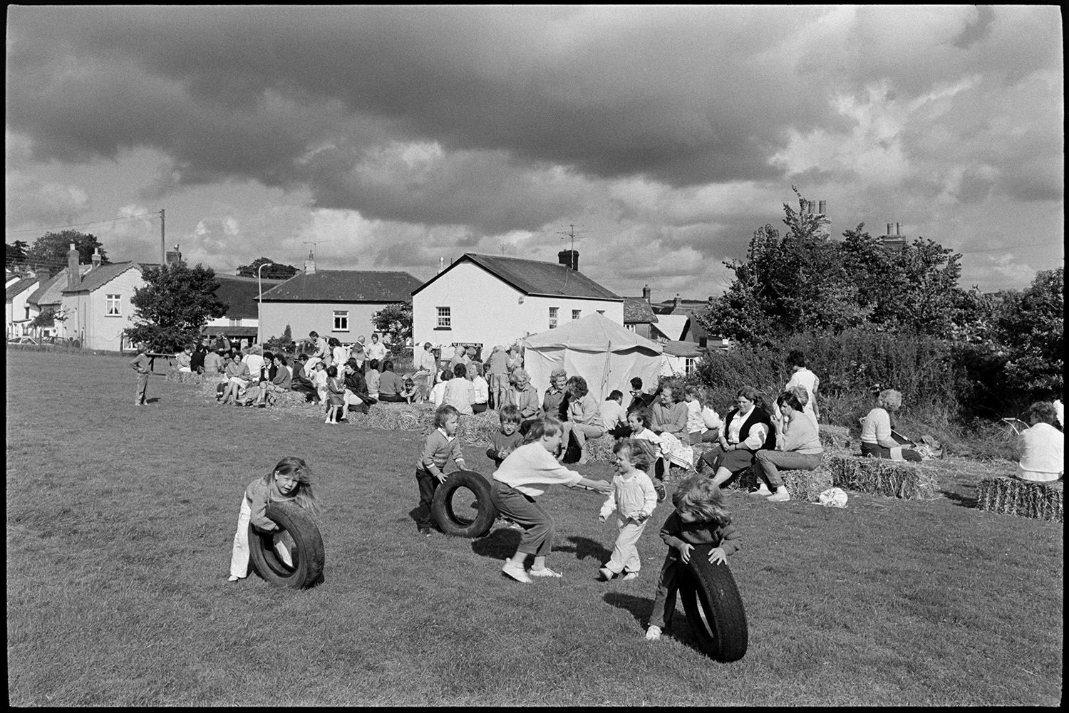 Sports Day, parachute landing races and spectators. 
[Children playing with tyres at Dolton School sports day. Men and woman are sat on hay bales watching and a refreshment tent can be seen in the background.]