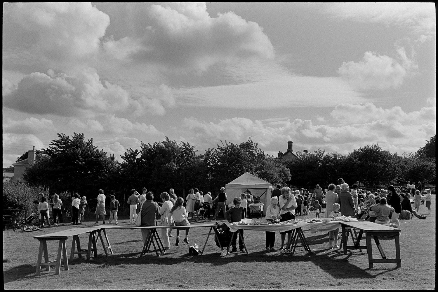 Sports Day, parachute landing races and spectators. 
[Men, woman and children at Dolton School sports day. Some people are helping themselves to cakes laid out on trestle tables and a refreshment tent is visible in the background.]