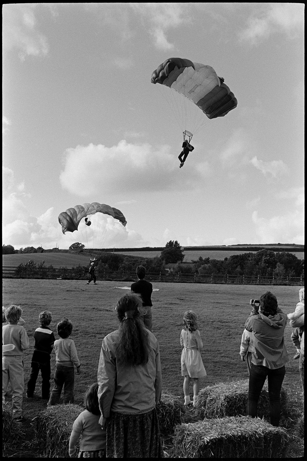 Sports Day, parachute landing races and spectators. 
[Two parachutists landing on the sports field at Dolton School sports day. Children and adults are watching them.]