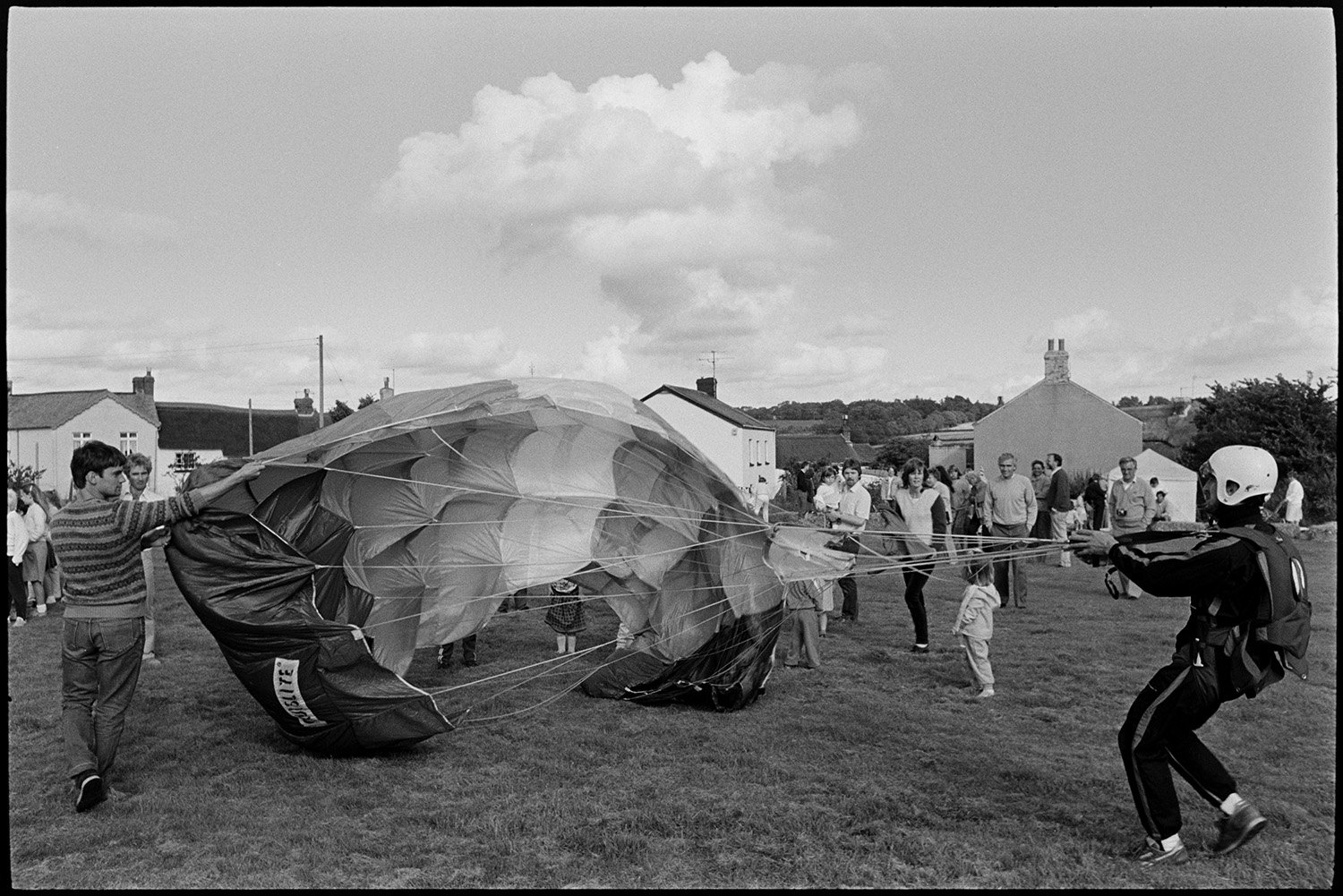 Sports Day, parachute landing races and spectators. 
[A man helping a parachutist with their parachute who has just landed in the field at Dolton School sports day. Children and adults are watching in the background.]