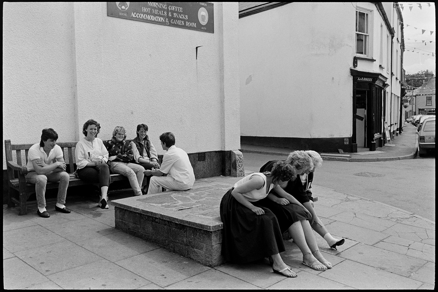 People sitting in middle of town chatting. 
[Young men and women sitting on a bench and concrete plinth outside the Red Lion pub in Chulmleigh. A&R Rogers Chemist can be seen across the street, which is decorated with bunting.]
