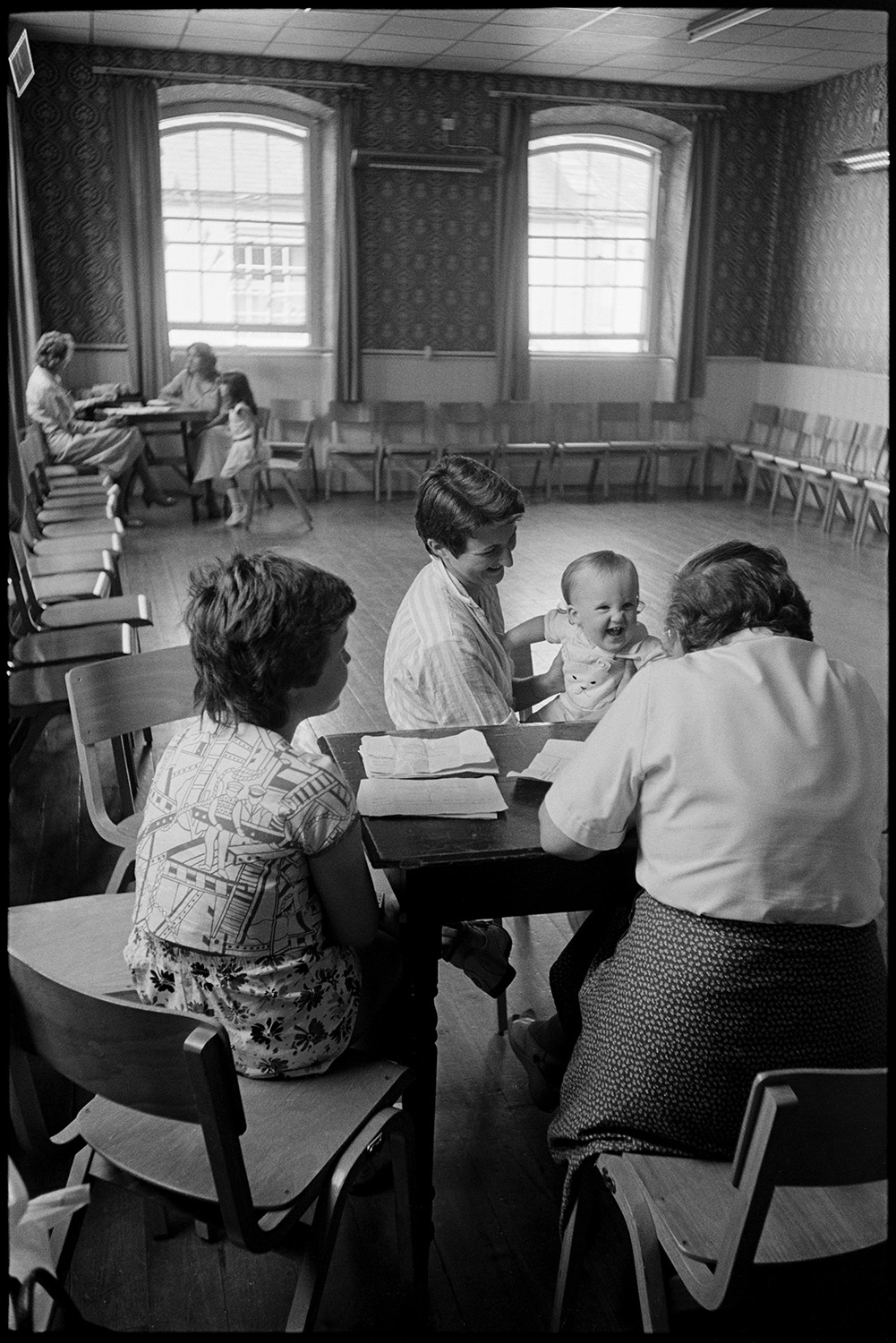 Fair. Baby competition, woman judging. Mothers and babies waiting to be judged. 
[Two women judging a baby with its mother at the baby competition for Chulmleigh Fair in Chulmleigh Town Hall. Seats are positioned around the hall and two other women and a child can be seen in the background.]