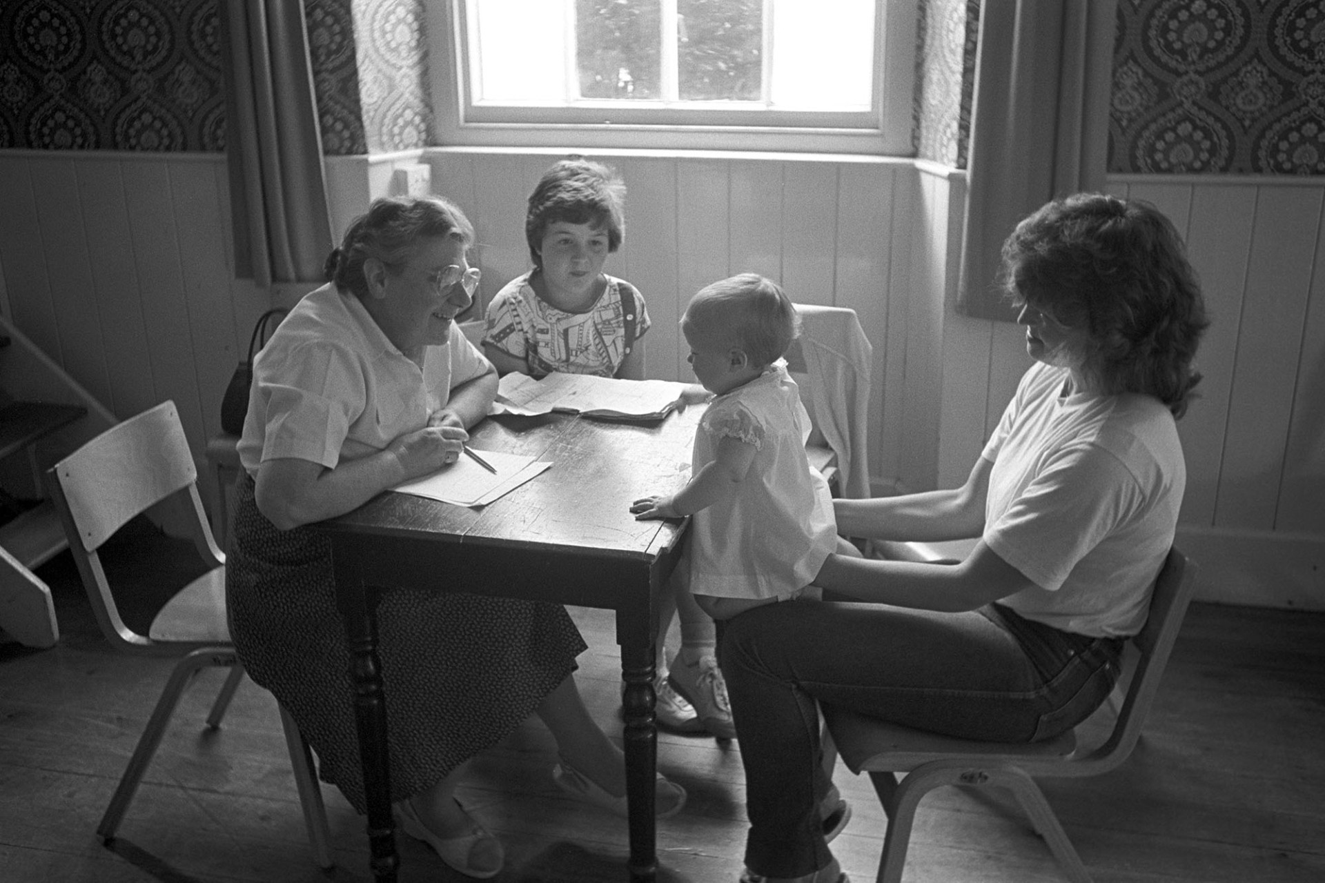 Fair, Baby competition, woman judging baby held by mother. 
[Sister Irene Morris judging a baby sat on its mother's lap in the baby competition at Chulmleigh Fair in Chulmleigh Town Hall.]