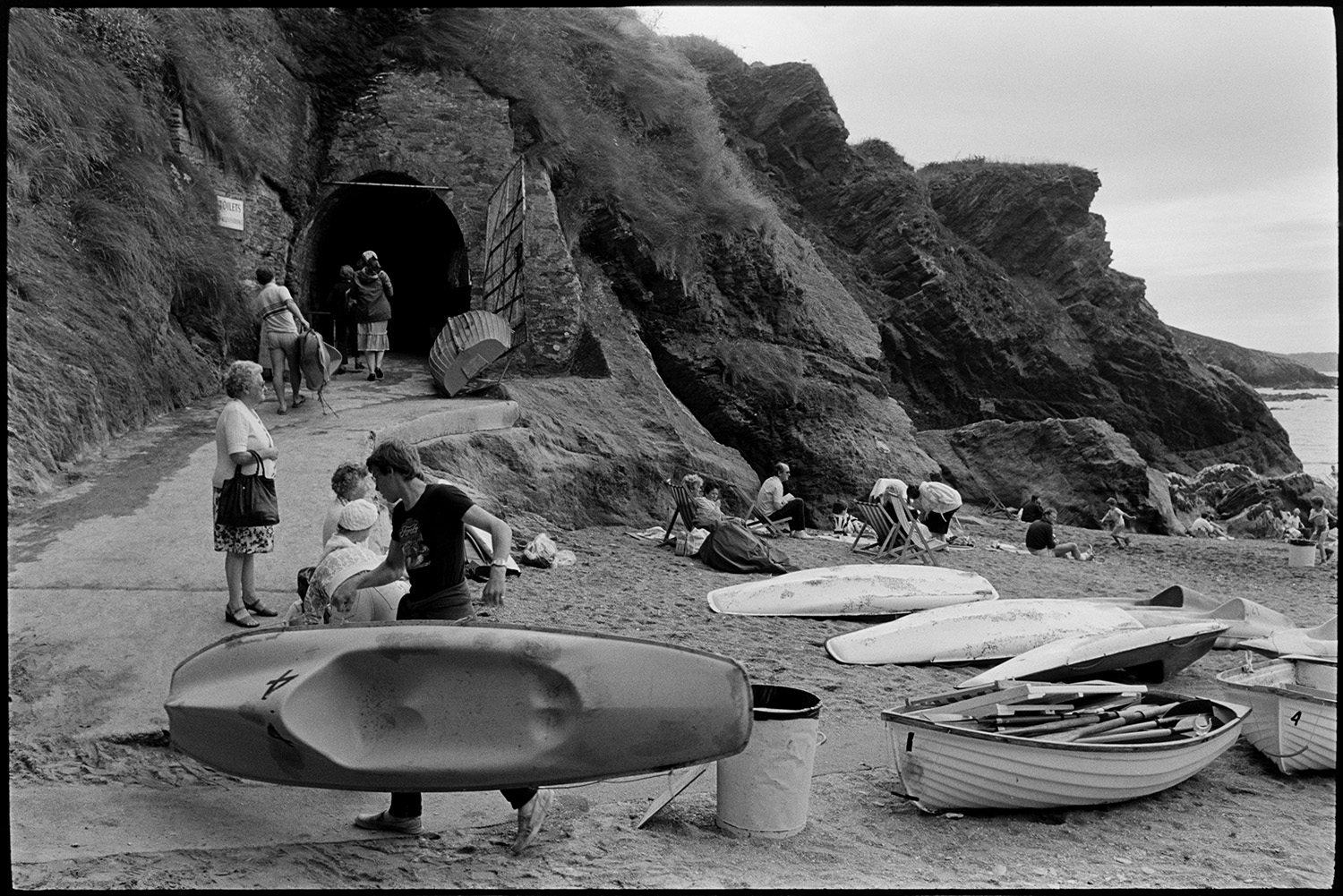 Entrance to tunnel and rocky beach with shop. People climbing on rocks. Boats. 
[People on Ilfracombe beach. Some are sat in deckchairs next to canoes and a rowing boat with oars on the beach. A man is carrying a small boat up a concrete path to a tunnel cut through the cliff to the beach.]