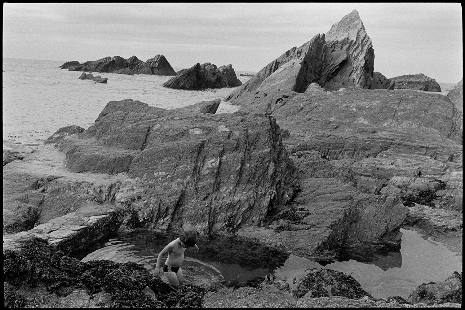 Entrance to tunnel and rocky beach with shop. People climbing on rocks. Boats. 
[A child exploring rock pools on a rocky outcrop from the cliffs at Ilfracombe beach.]