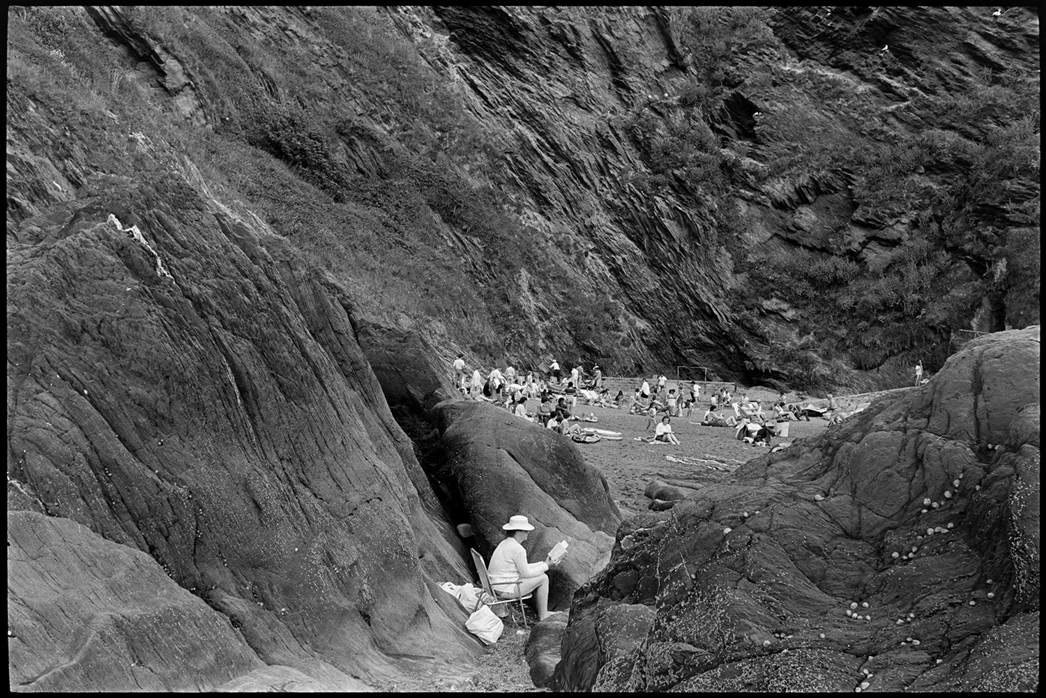 Entrance to tunnel and rocky beach with shop. People climbing on rocks. Boats. 
[People sitting and sun bathing on the beach at Ilfracombe, with cliffs in the background. A person is sat on a garden chair reading a book, at the bottom of the cliff in the foreground.]