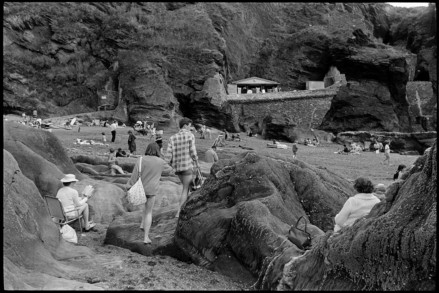 Entrance to tunnel and rocky beach with shop. People climbing on rocks. Boats. 
[A man and woman walking along rocks at Ilfracombe beach. Two people are sat by the rocks at the bottom of the cliff in the foreground. More people can be seen sitting and sun bathing on the beach and a hut selling beach goods is visible in the background.]