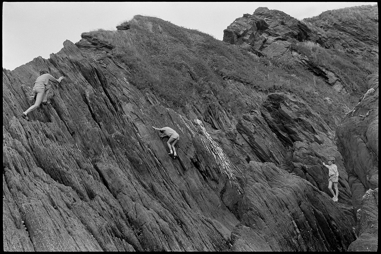 Entrance to tunnel and rocky beach with shop. People climbing on rocks. Boats. 
[Three children climbing on a rocky outcrop from the cliffs at Ilfracombe.]