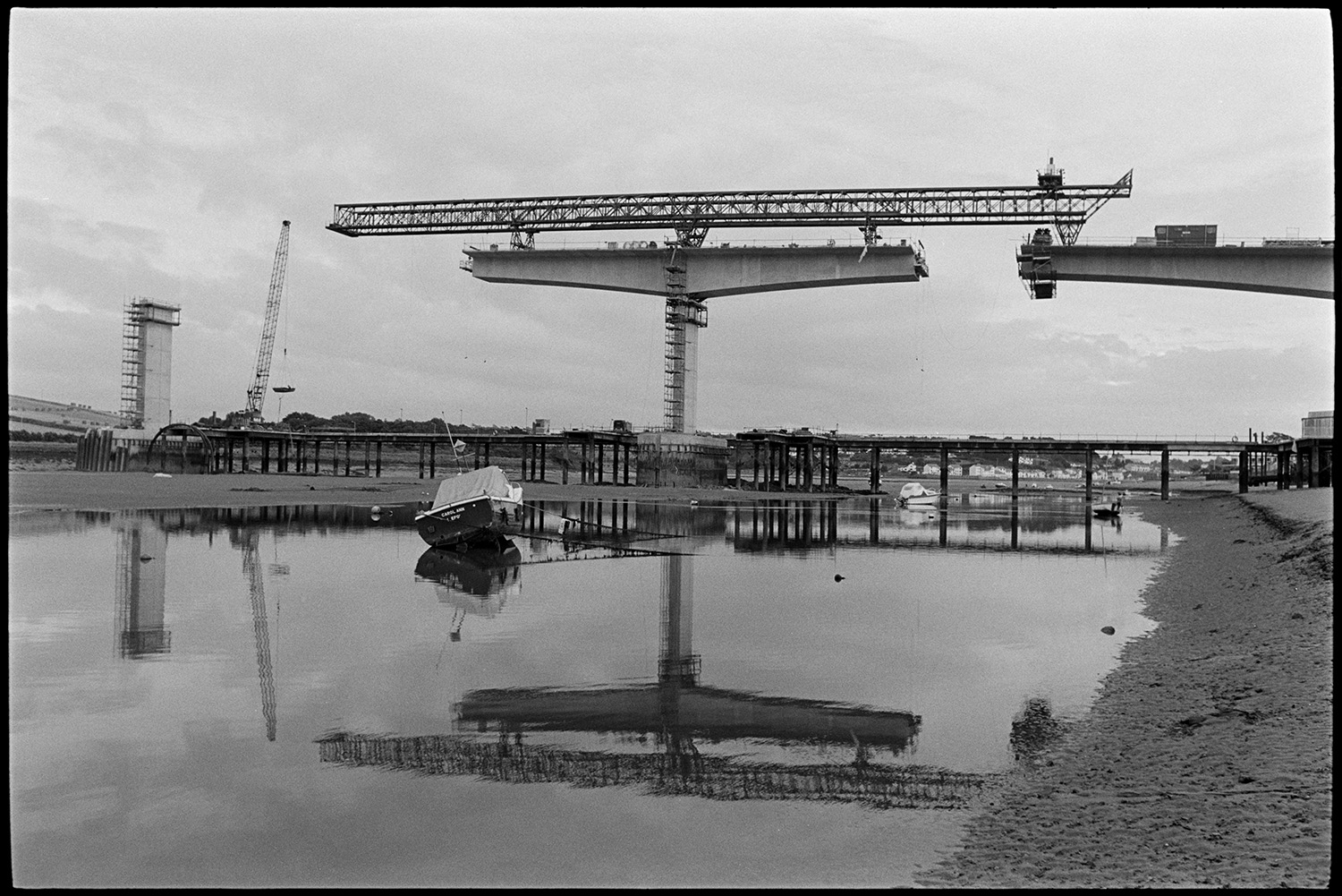 New bridge under construction over river, mudflats with small craft, boats. 
[Cranes constructing Bideford New Bridge across the River Torridge. Small boats can be seen moored on the river in the foreground and reflections of the cranes are visible in the river.]