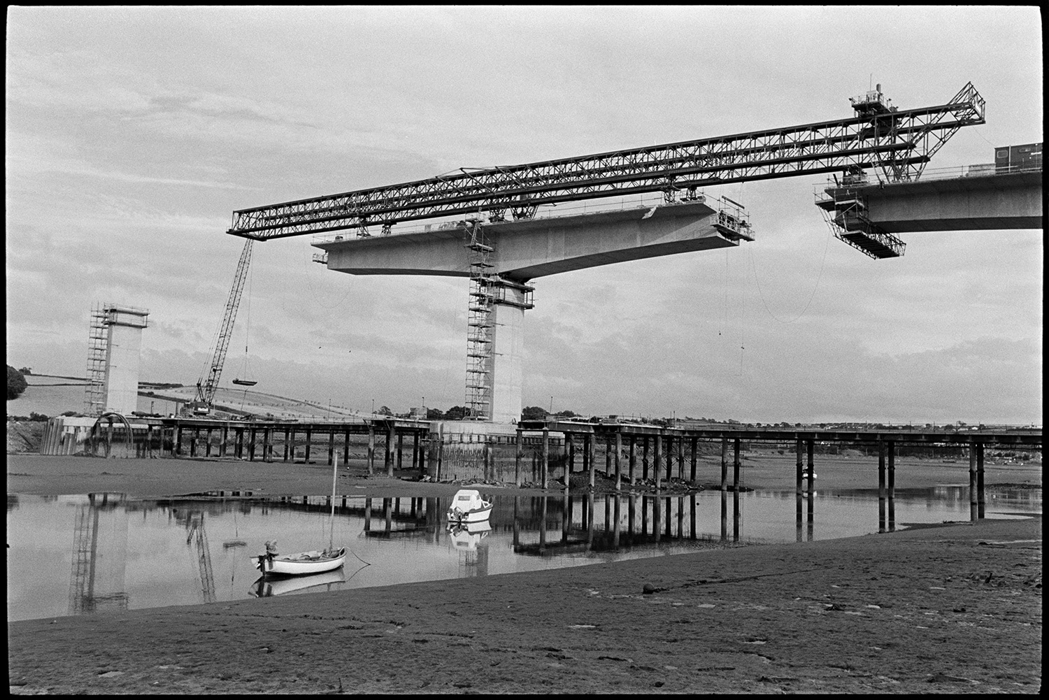 New bridge under construction over river, mudflats with small craft, boats. 
[Cranes constructing Bideford New Bridge across the River Torridge. Small boats can be seen moored on the river in the foreground.]