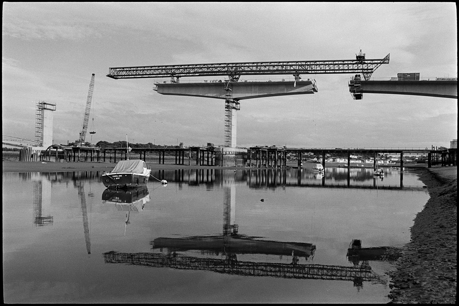 New bridge under construction over river, mudflats with small craft, boats. 
[Cranes constructing Bideford New Bridge across the River Torridge. Small boats can be seen moored on the river in the foreground and reflections of the cranes are visible in the river.]