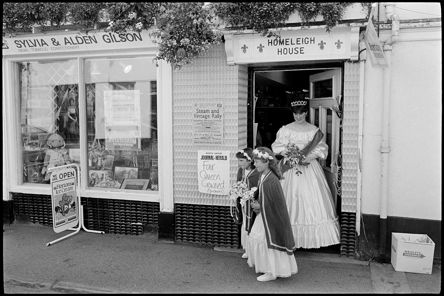 Fair Queen and attendants in street outside newsagents shop and in town hall. White dress. 
[Emma Bending, the Chulmleigh Fair Queen coming out of a newsagents shop with two attendants, at Homeleigh House in Chulmleigh. She is wearing a white dress and a sash. The names Sylvia & Alden Gilson are above the shop window. A doll and framed pictures are displayed in the window. Two posters are pinned up outside the hop doorway, including an advert for a Steam and Vintage Rally.]