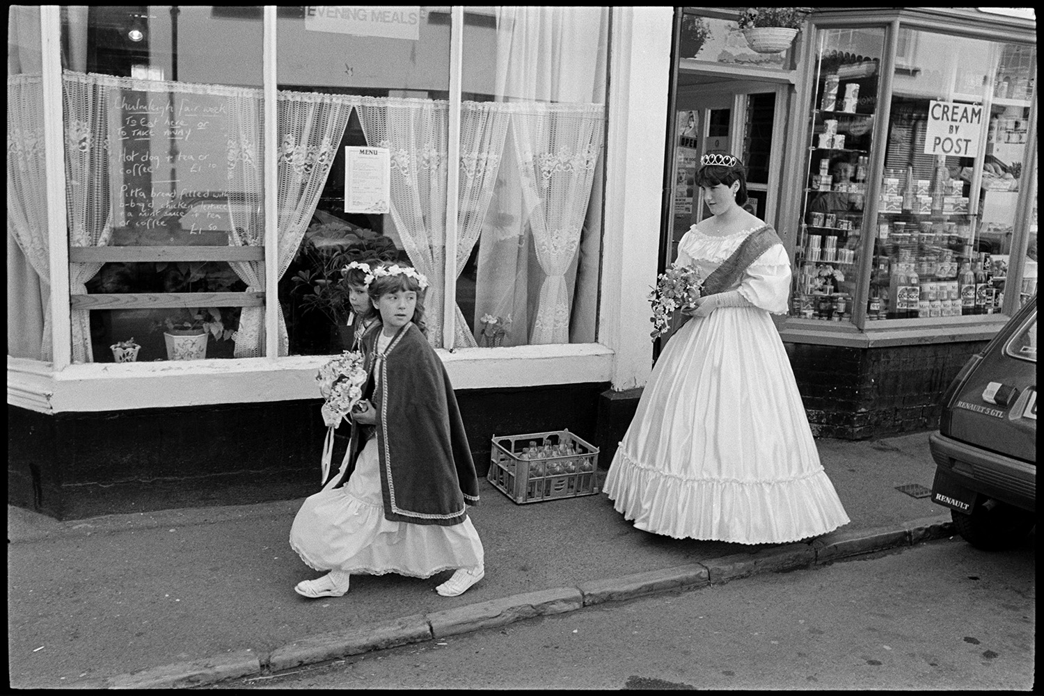 Fair Queen and attendants in street outside newsagents shop and in town hall. White dress. 
[Emma Bending, the Chulmleigh Fair Queen walking along a street with two attendants in Chulmleigh. She is wearing a white dress and a sash. They are passing a shop front displaying canned food and a café window. A crate of empty milk bottles is on the pavement.