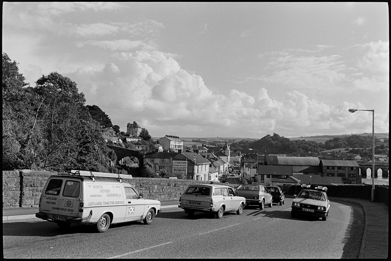 View over town with cars parked beside road. 
[A view of East the Water, Bideford with buildings and cars on a road. Part of Bideford Long Bridge, also known as Bideford Old Bridge, can be seen in the background. In the distance trees, fields and clouds are visible.]
