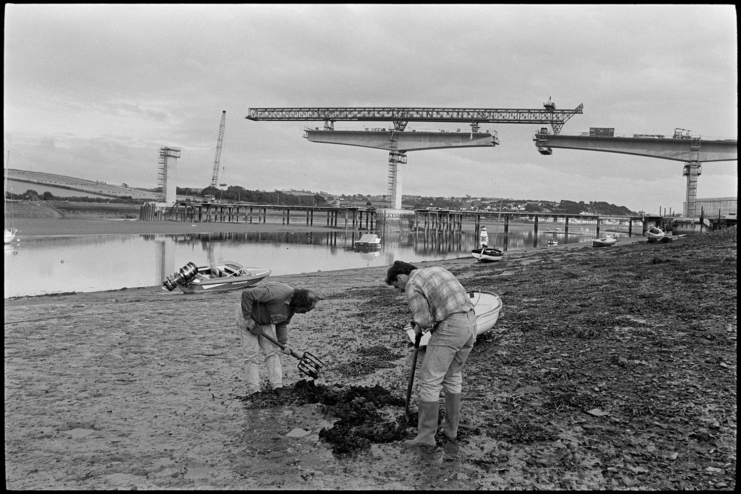 River views with new bridge being built, men digging worms for bait, boats. Garden chairs. 
[Two men digging for worms for bait on the mud flats of the river Torridge at Bideford. Cranes are constructing Bideford New Bridge in the background. Small boats are moored on the river and the mud flats.]