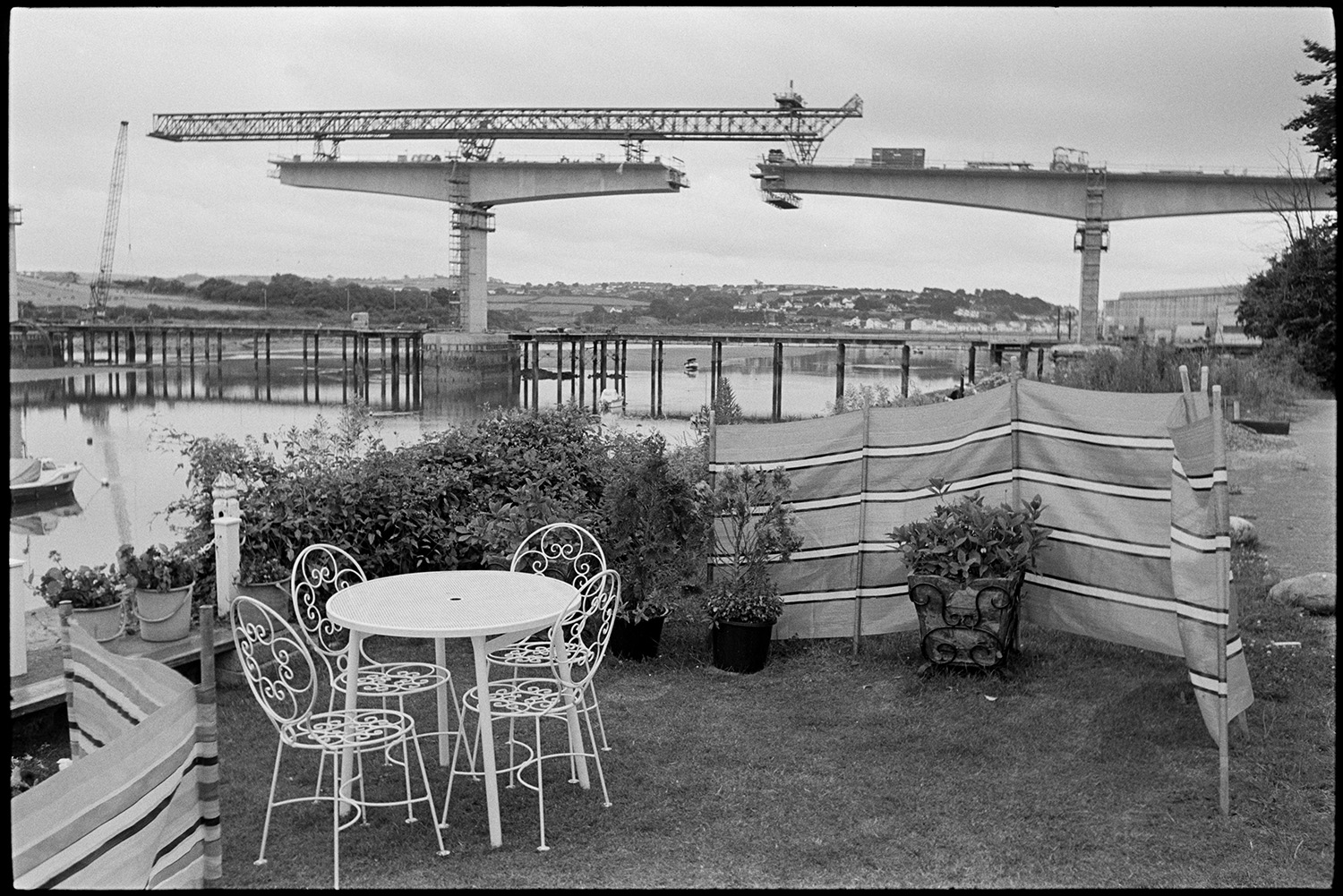 River views with new bridge being built, men digging worms for bait, boats. Garden chairs. 
[A garden with wind breaks, plant pots and a garden table and chairs by the River Torridge at Bideford. Cranes can be seen constructing Bideford New Bridge in the background, and a few boats are on the river.]