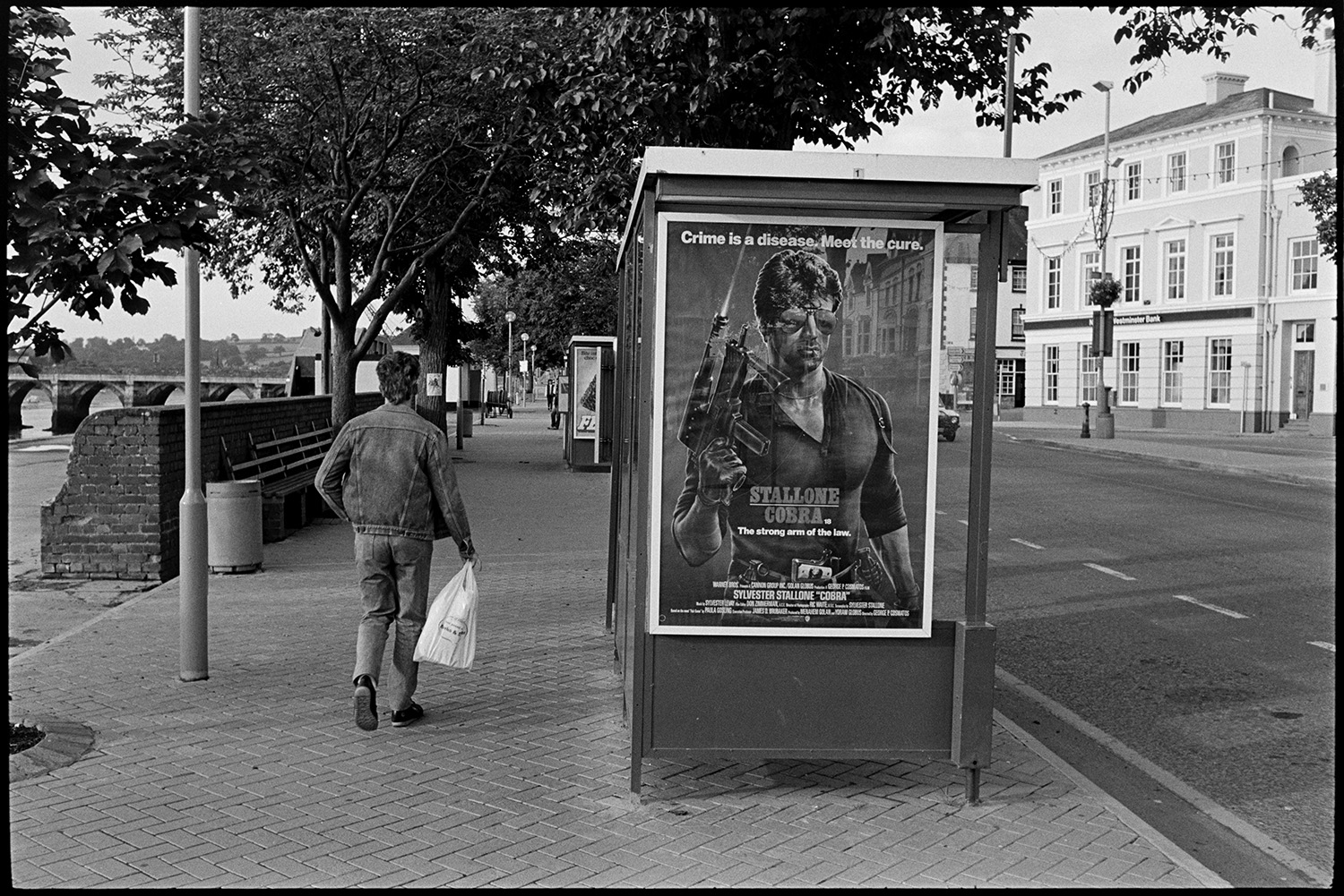 Cinema film advert on the end of bus shelter for violent macho Stallone Cobra - sad! 
[A person holding a carrier bag walking past a bus shelter, with an advert for the film Cobra, starring Sylvester Stallone, alongside the riverfront at Bideford. Bideford Long Bridge, also known as Bideford Old Bridge, can be seen in the background.]