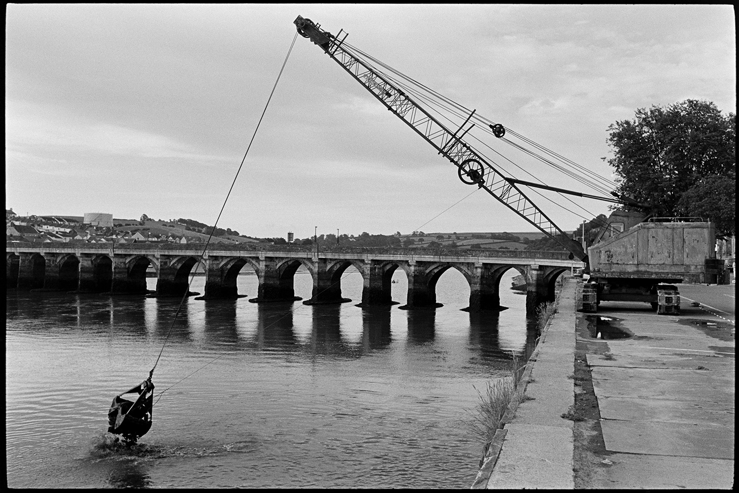 Crane and grab dredger working off quay, old bridge behind. 
[A crane and mechanical grab dredging off Bideford Quay. Bideford Long Bridge, also known as Bideford Old Bridge, can be seen in the background.]