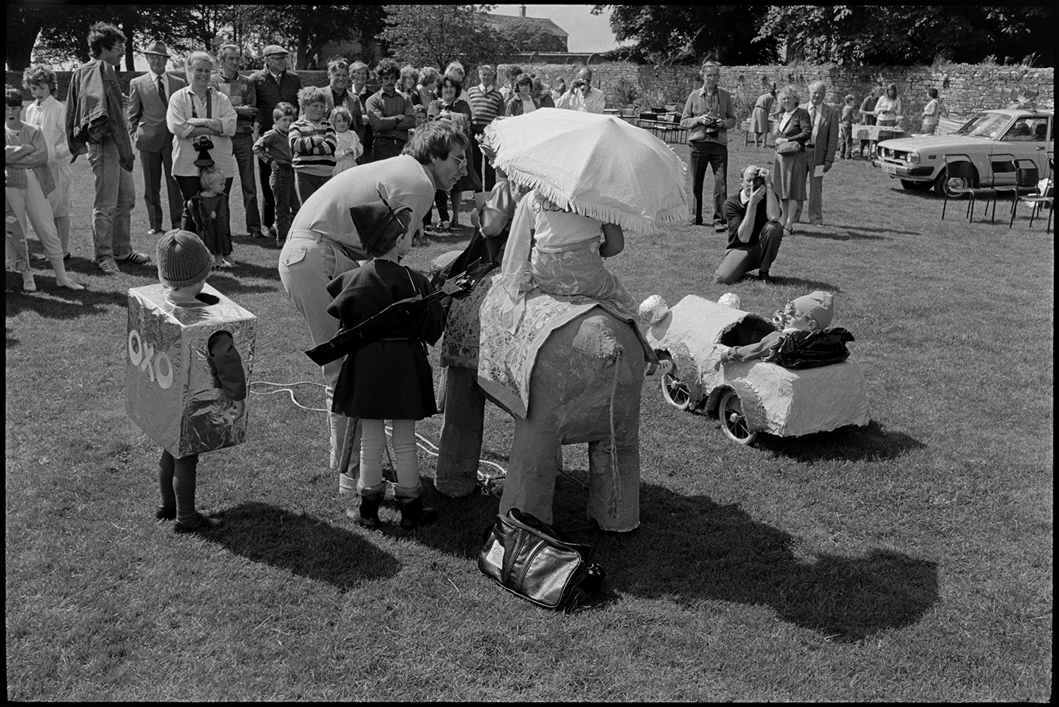 Village revel, fete, band, dance on green, stalls, jumble, guess weight of the Ram, fancy dress.
[People watching children lined up in fancy dress at Beaford Revel on the village green. One child is sat on a model elephant, another is dressed as an Oxo cube and one is Noddy sat in a model car.]