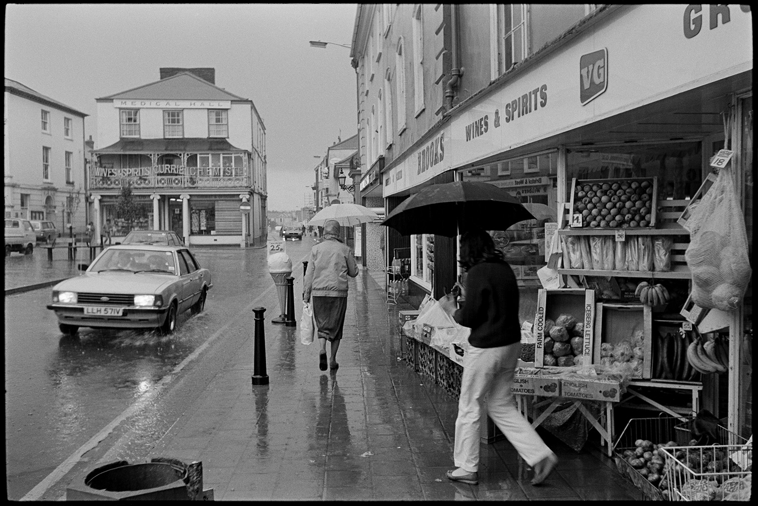 Street scene in heavy rain, umbrellas, shop fronts.
[South Molton square in heavy rain. Pedestrians with umbrellas are walking on the pavement alongside a car splashing through a puddle on the road. Brooks shop front with boxes of fruit and vegetables on display is visible and the Medical Hall with Currie's chemists shop is in the background.]