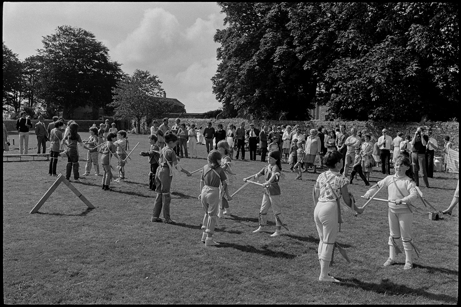 Village revel, fete, folk dance on green, stalls, jumble, guess weight of the Ram, fancy dress.
[Children Morris dancing at Beaford Revel on the village green. The churchyard wall with large trees and a crowd of spectator are visible in the background.]