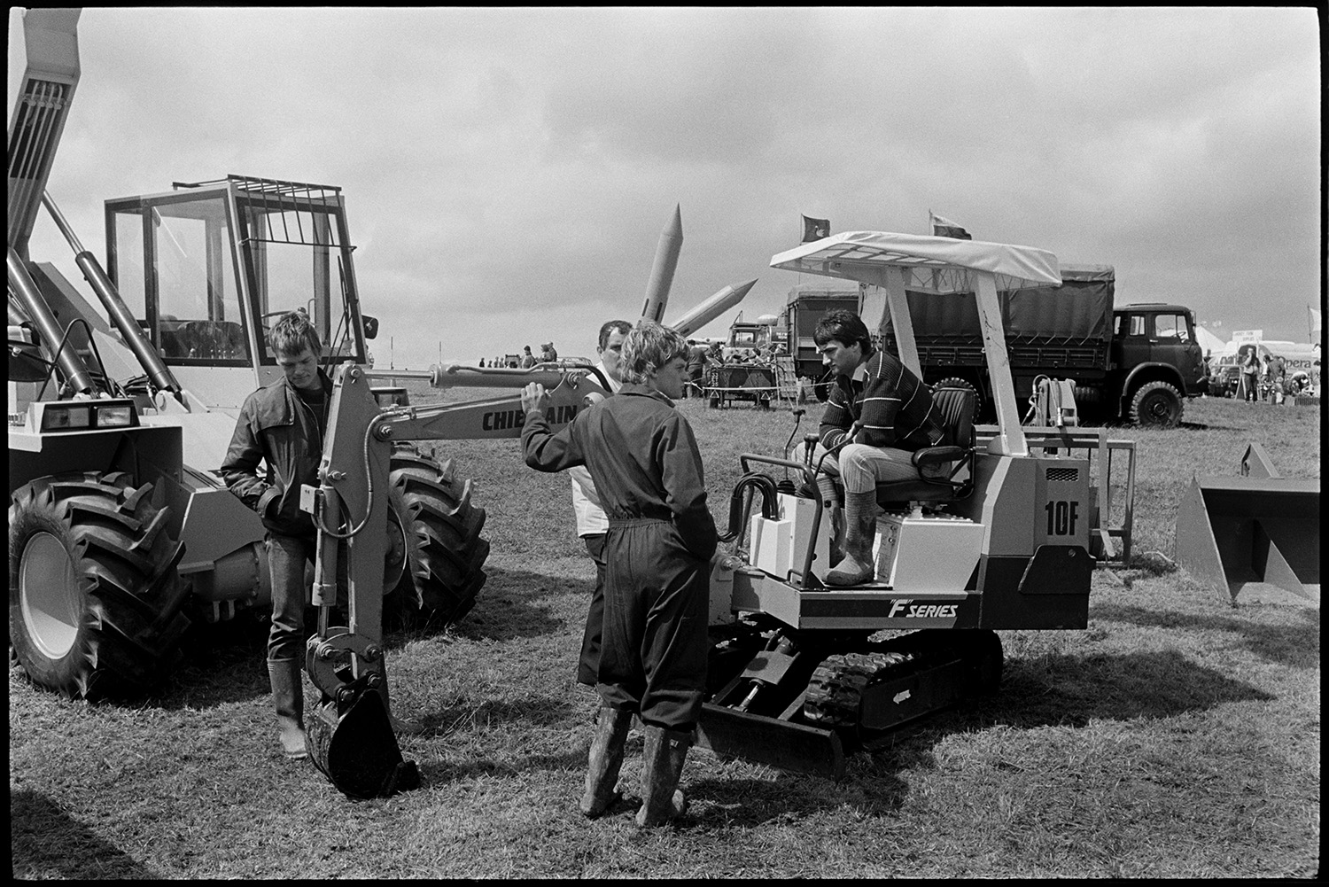 Agricultural show, judging sheep. Prize cattle in pens, spectators.
[Agricultural machinery on display at the North Devon Show near Alverdiscott. Four men are looking at a digger. Lorries, tents and spectators are visible in the background.]
