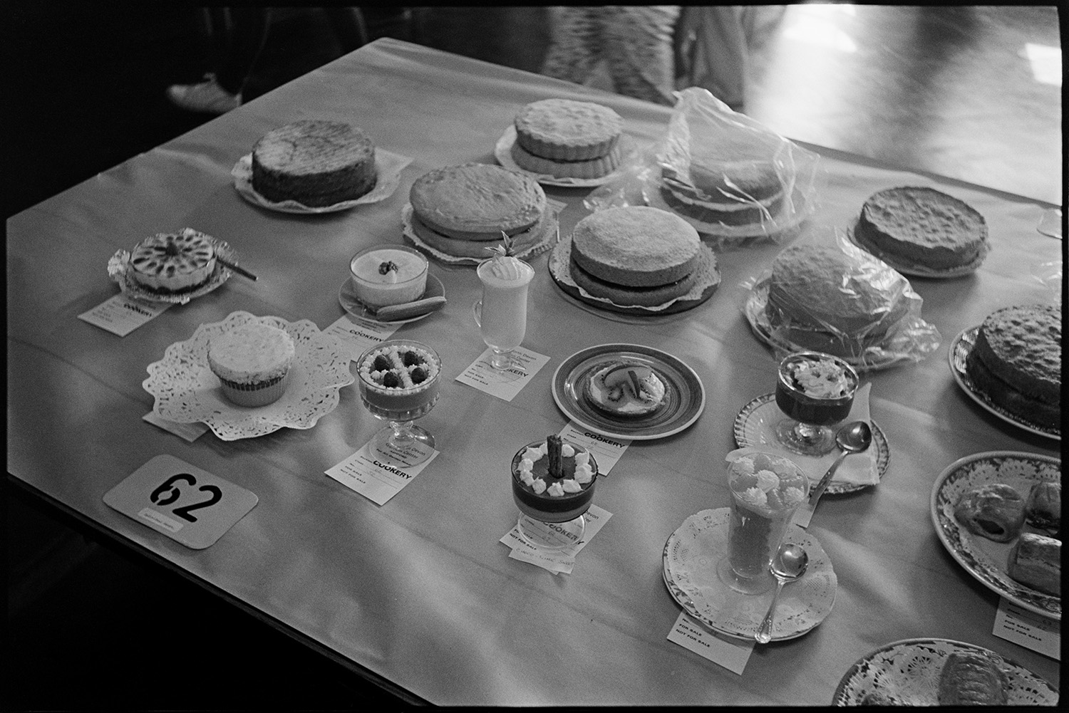 Flower show, displays of roses, cakes, vegetables, skittle alley.
[Display of cakes and desserts on a table at the Dolton Flower Show, in the village hall, for the cookery competition. A range of sponge cakes, various dessert dishes, and sausage rolls are displayed.]