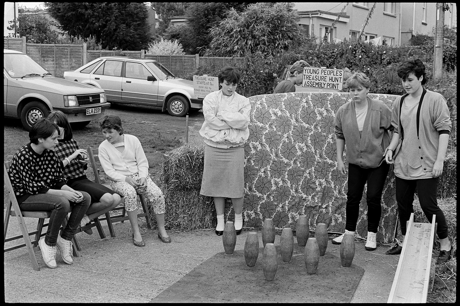 Flower show, displays of roses, cakes, vegetables, skittle alley.
[Teenage girls standing around the outdoor skittle alley at Dolton Flower Show. A mattress and straw bales are behind the skittles. Parked cars, signs for a Treasure Hunt, shrubs and houses are visible in the background.]