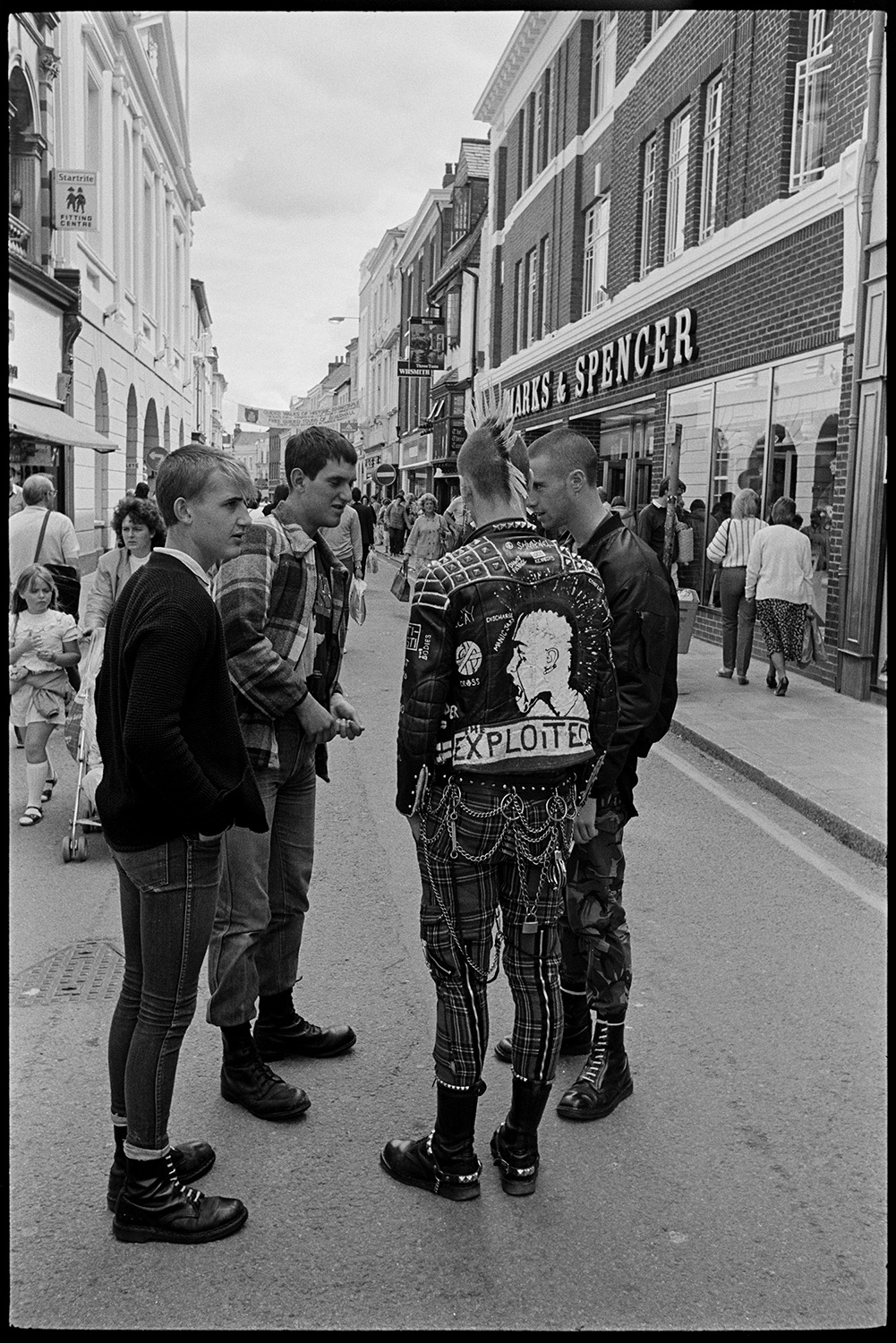 Three (very nice) skinheads or punks in town, one with mohican haircut. Good clothes.
[Four young men, or punks, standing in Barnstaple High Street. One is wearing tartan trousers, a leather jacket and a mohican haircut. Marks and Spencer shop front is visible in the background.]