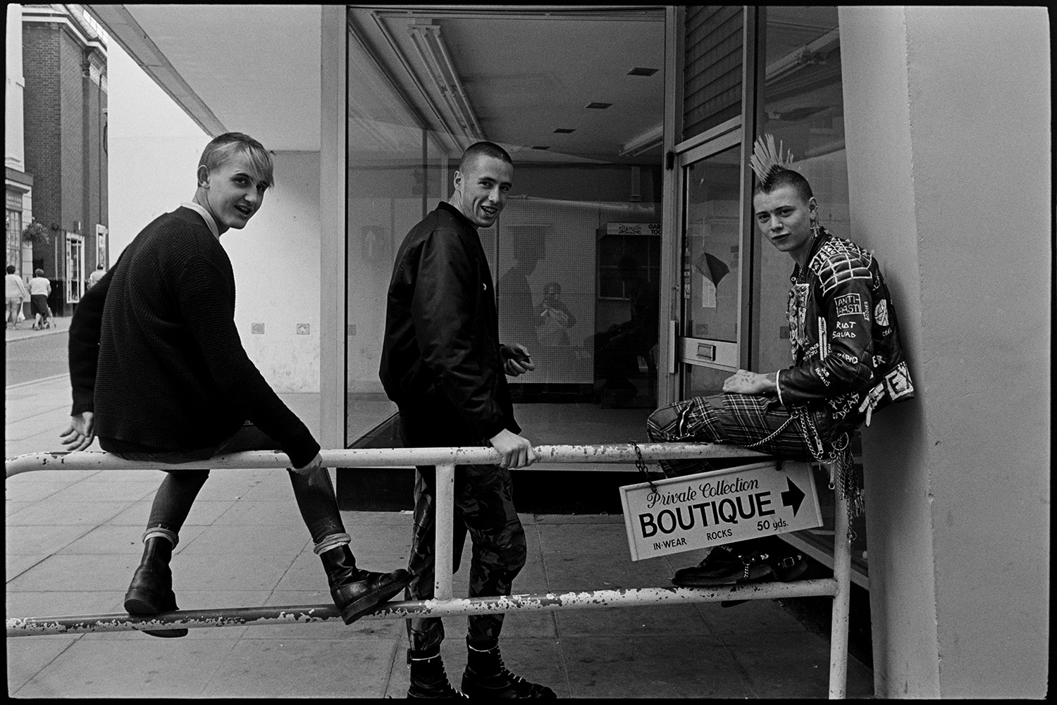 Three (very nice) skinheads or punks in town, one with mohican haircut. Good clothes.
[Three young men, or punks, outside an empty shop in Barnstaple High Street. One is wearing tartan trousers, a leather jacket and a mohican haircut. They are all wearing Dr Marten boots.]