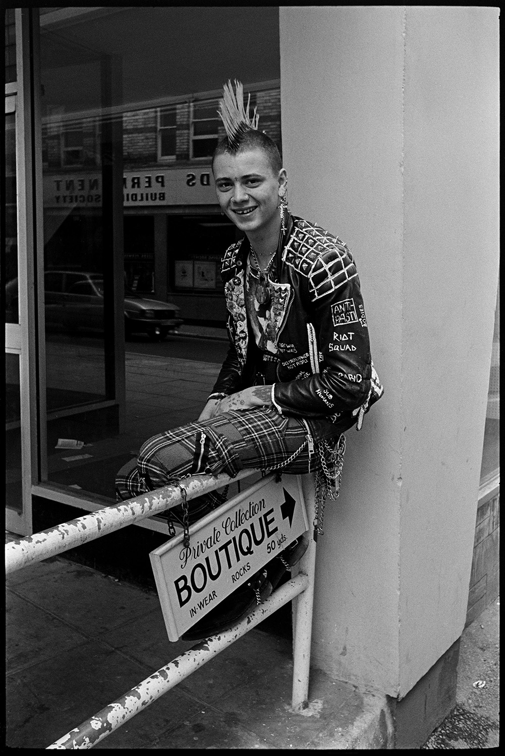 Three (very nice) skinheads or punks in town, one with mohican haircut. Good clothes.
[A young man, or punk, dressed in tartan trousers, a leather jacket, and with a mohican haircut, sitting on a railing outside an empty shop window on Barnstaple High Street. A small sign for a boutique hangs on the railings.]