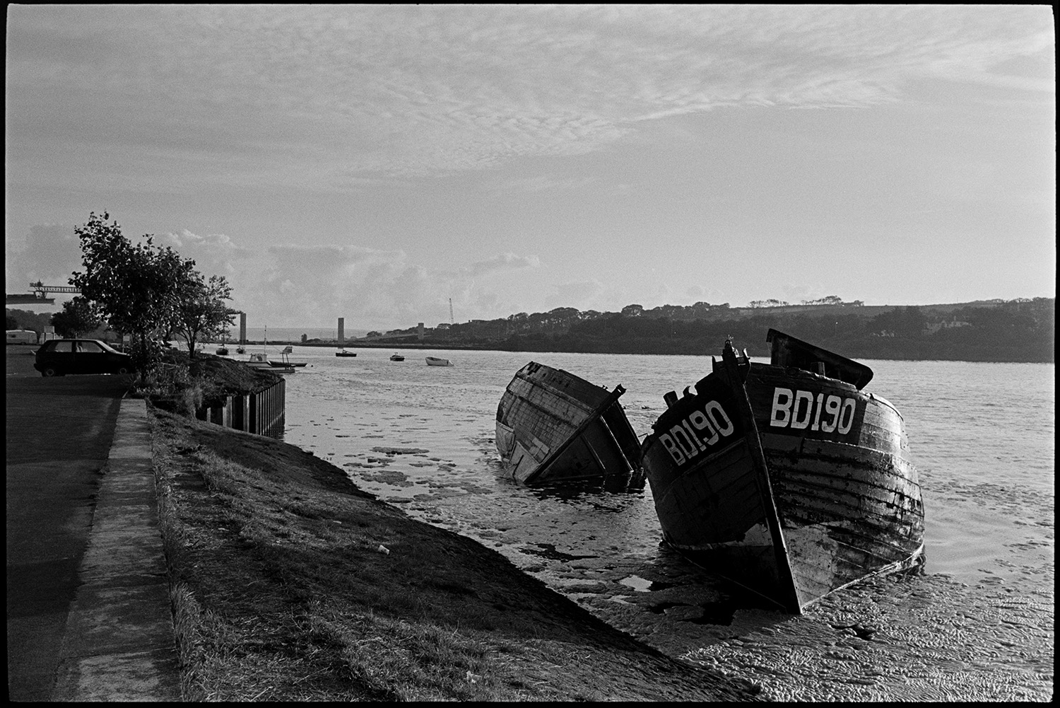 Abandoned fishing boats, bridge construction in background, early morning.
[Two abandoned wooden fishing boats near the river bank at Bideford on the River Torridge. Boats on the river and the pillars for Bideford New Bridge are under construction in the background. The boat in the foreground has the number 'BD190' painted on it.]