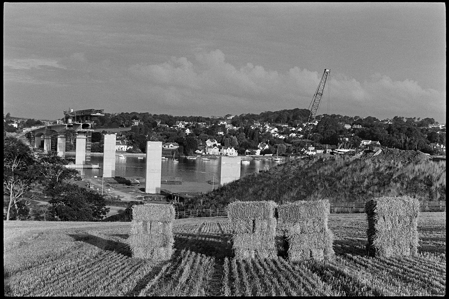 Bales of hay with bridge under construction in background.
[Straw bales standing in a field at Bideford with Bideford New Bridge being built over the River Torridge in the background.]